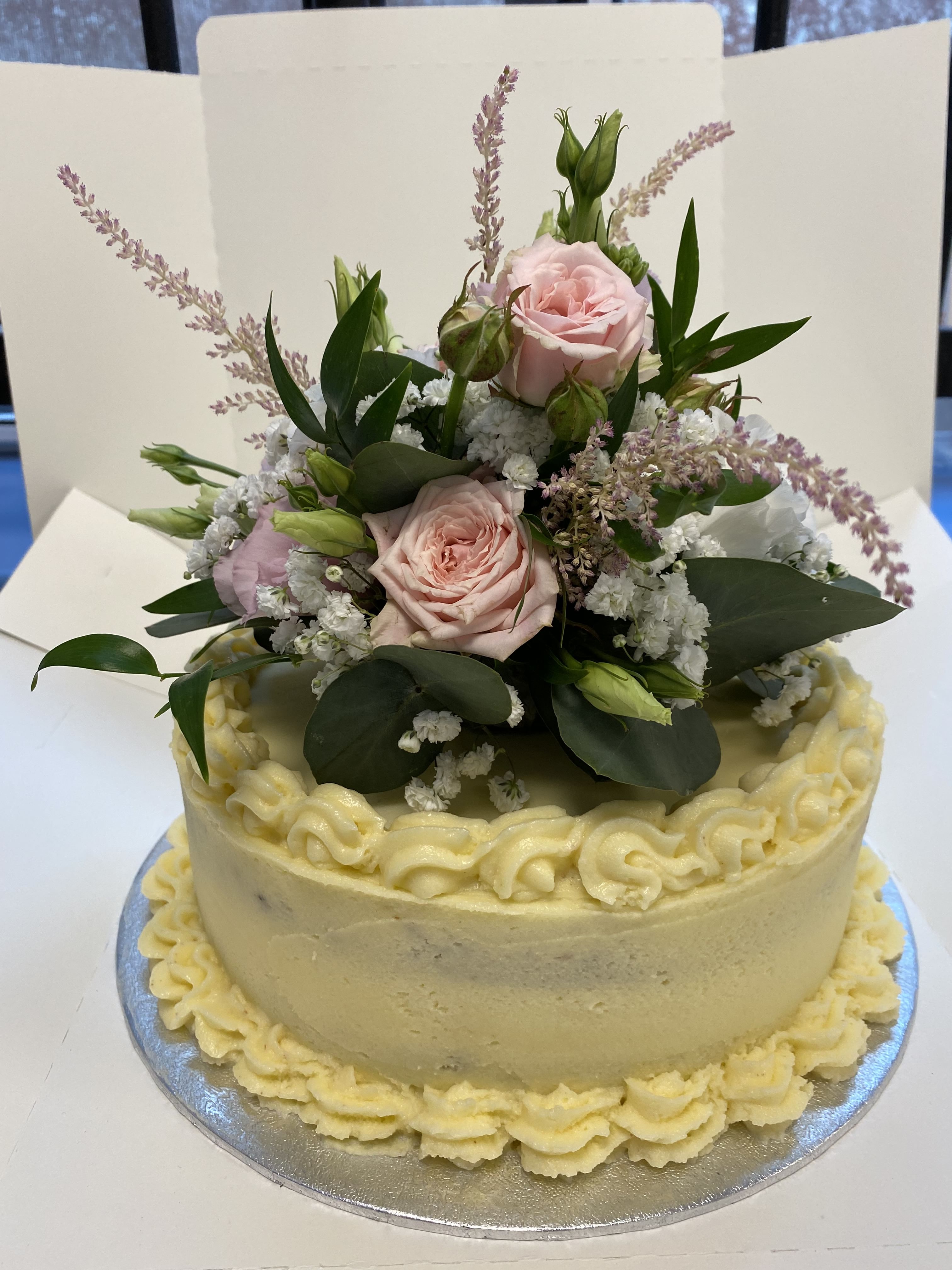A beautifully decorated cake with intricate yellow frosting edging. The top of the cake is adorned with a bouquet of pink and white roses, green leaves, and sprigs of delicate white flowers. The cake is displayed against a plain background.